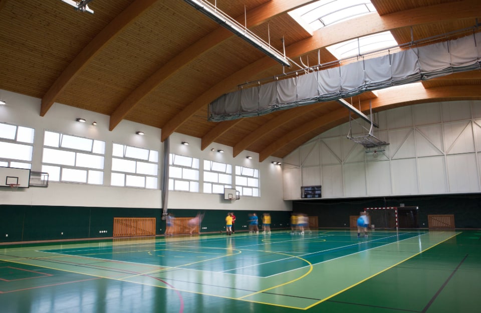 Campo sportivo indoor multifunzionale con tetto in legno e alcuni giocatori sul campo, visti da lontano
