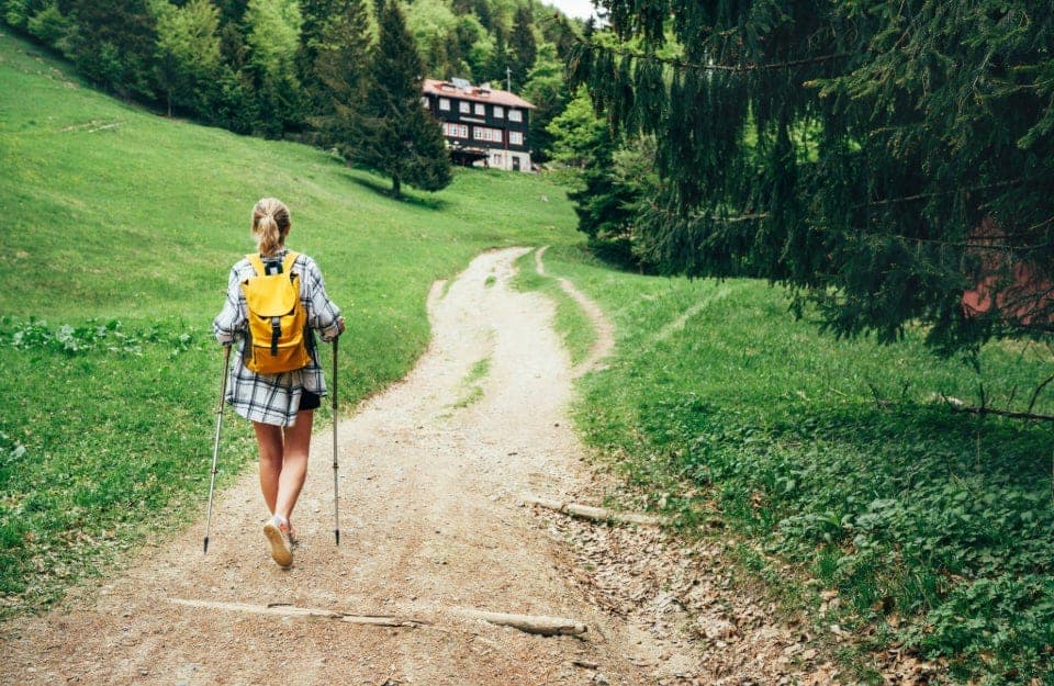 Una ragazza bionda in pantaloncini, vista di spalle, sta camminando con i bastoni da trekking lungo un sentiero che porta a un rifugio alpino tra i boschi