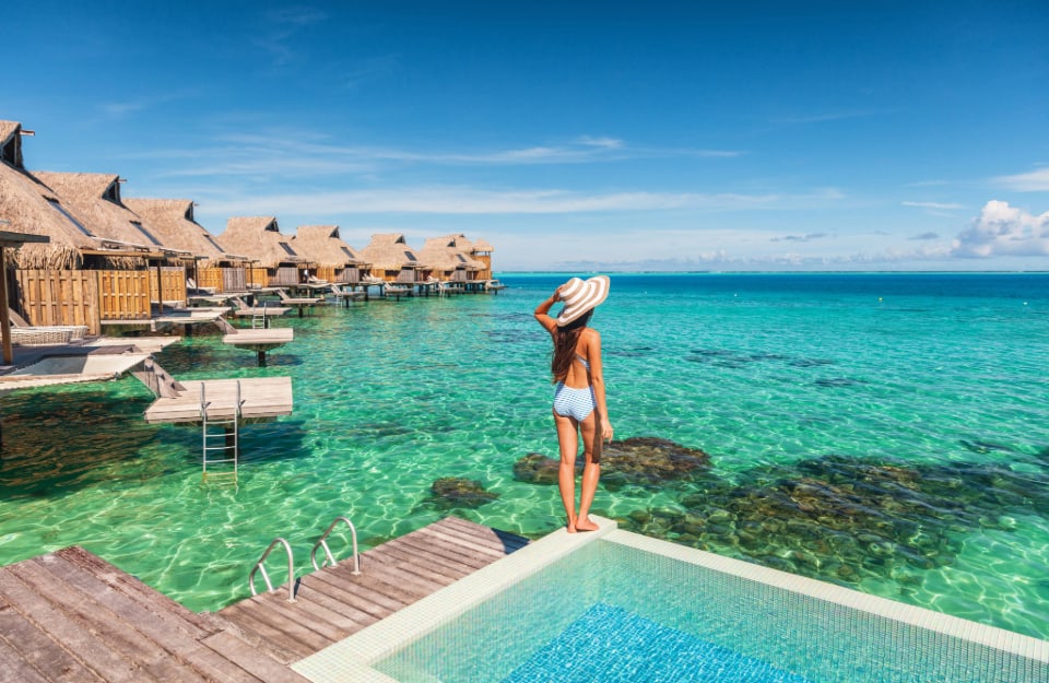 Una donna in costume con un grande cappello, vista di spalle in piedi sul bordo di una piscina di un villaggio turistico, con l'acqua cristallina del mare a pochi passi, e una fila di bungalow su palafitte