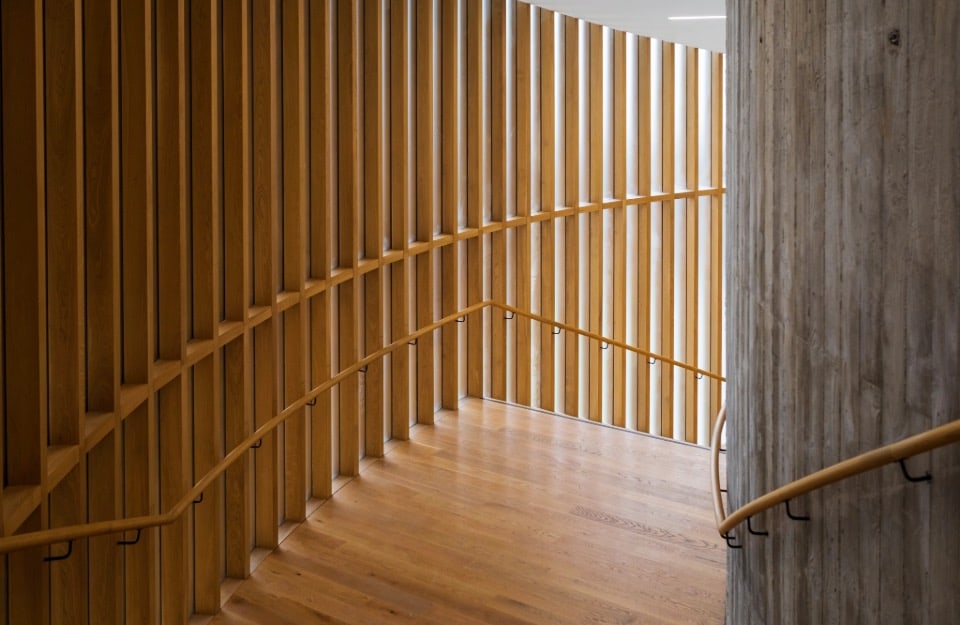 Uno spazio architettonico interno con una scala curva in legno dotata di corrimano su entrambi i lati. La scala è racchiusa da doghe verticali di legno su un lato e da un muro di cemento sull'altro. Le doghe in legno permettono alla luce di filtrare attraverso, creando un interessante gioco di luci e ombre sul pavimento e sulle pareti. Il pavimento e i corrimano sono realizzati nello stesso legno