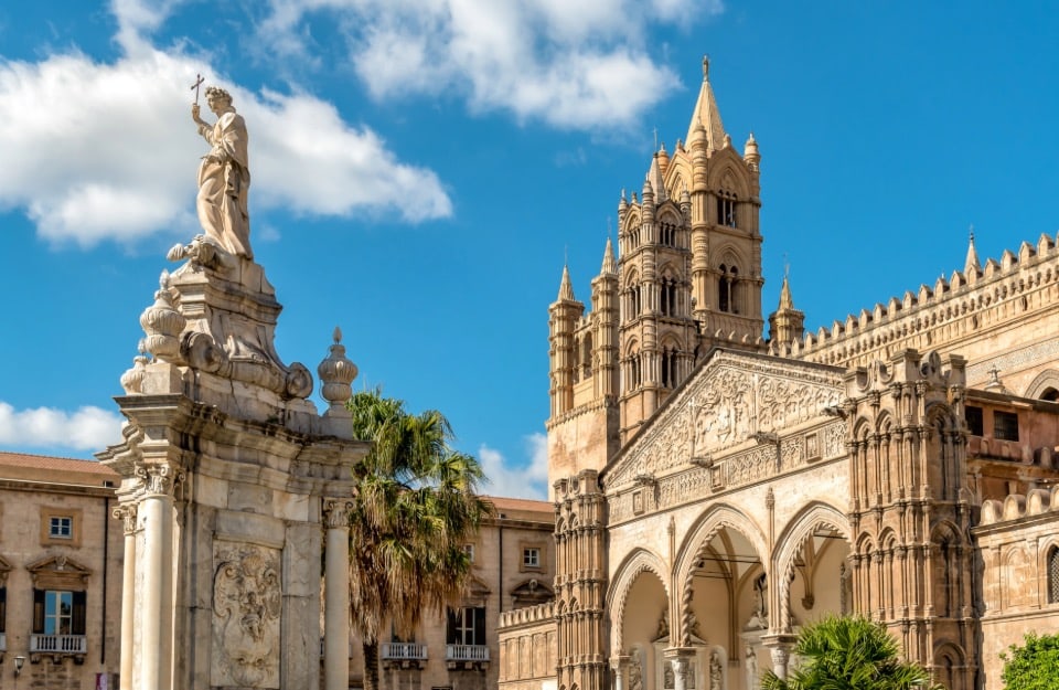 La Cattedrale di Palermo. In primo piano, c'è una statua monumentale di una figura religiosa che tiene una croce, posta su un piedistallo decorato con dettagli architettonici elaborati. Sullo sfondo, si può vedere la facciata della cattedrale, caratterizzata da archi gotici, intricate sculture e un campanile imponente. La cattedrale è un esempio significativo di architettura normanna e gotica