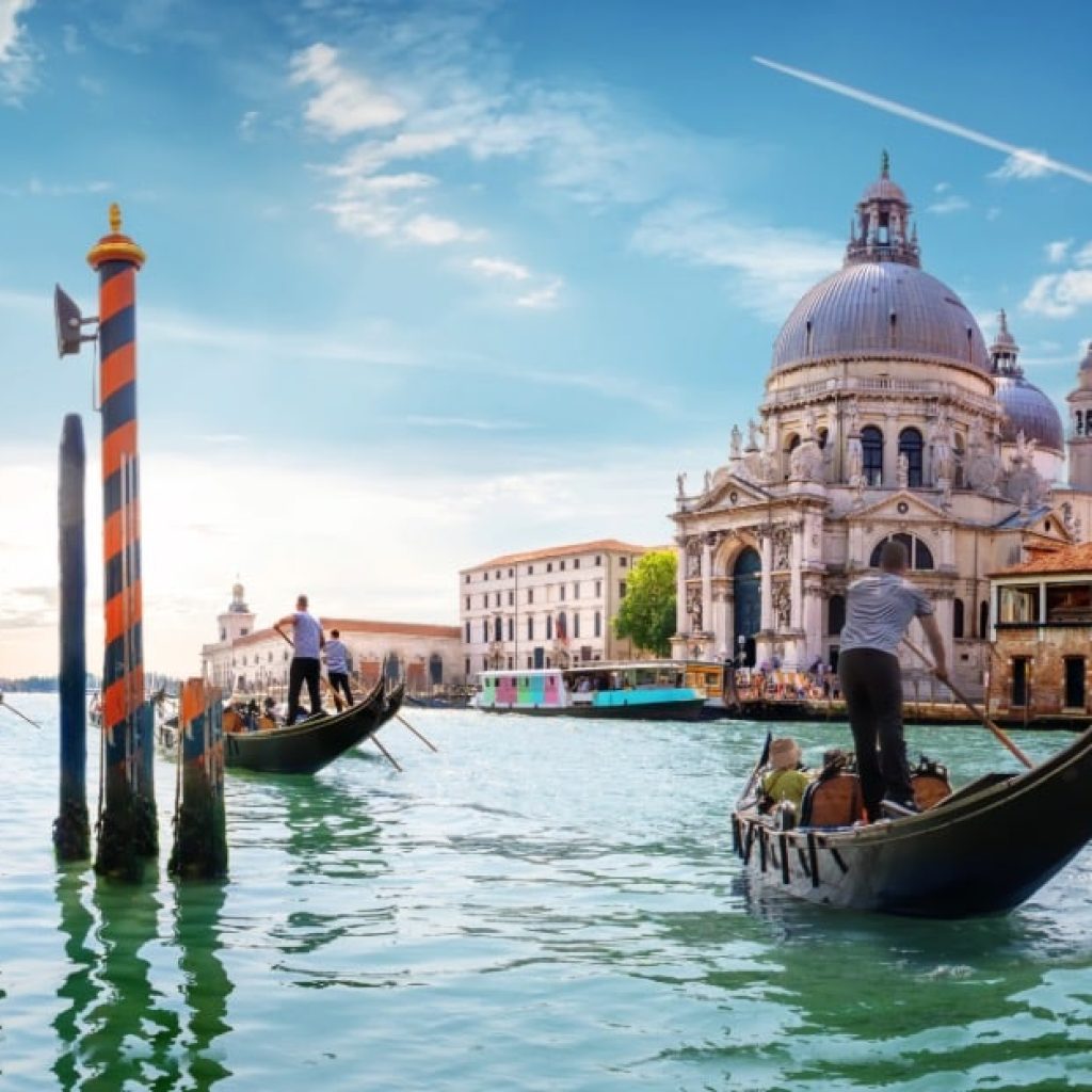 Una scena iconica di Venezia. In primo piano ci sono due gondole con gondolieri che remano attraverso un canale. Sullo sfondo si vede la Basilica di Santa Maria della Salute, un famoso edificio barocco con una grande cupola. L'acqua del canale è verde e riflette il cielo azzurro con alcune nuvole. Ci sono anche pali di ormeggio a strisce arancioni e nere che emergono dall'acqua